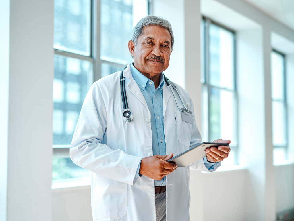 Portrait of senior male physician with digital tablet, looking at camera in hospital lounge